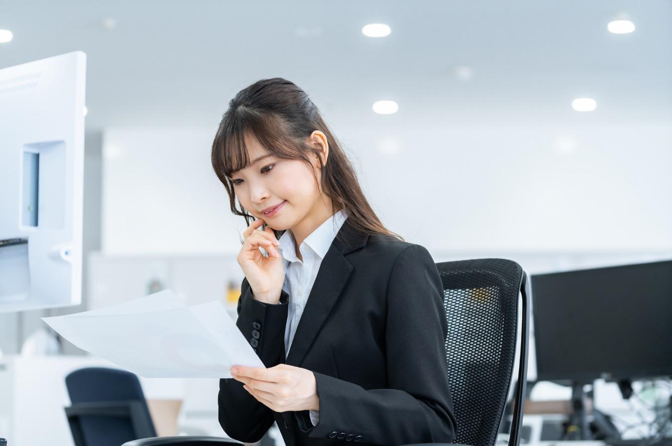 デスクトップPCを前に、手元の資料を見つめる女性