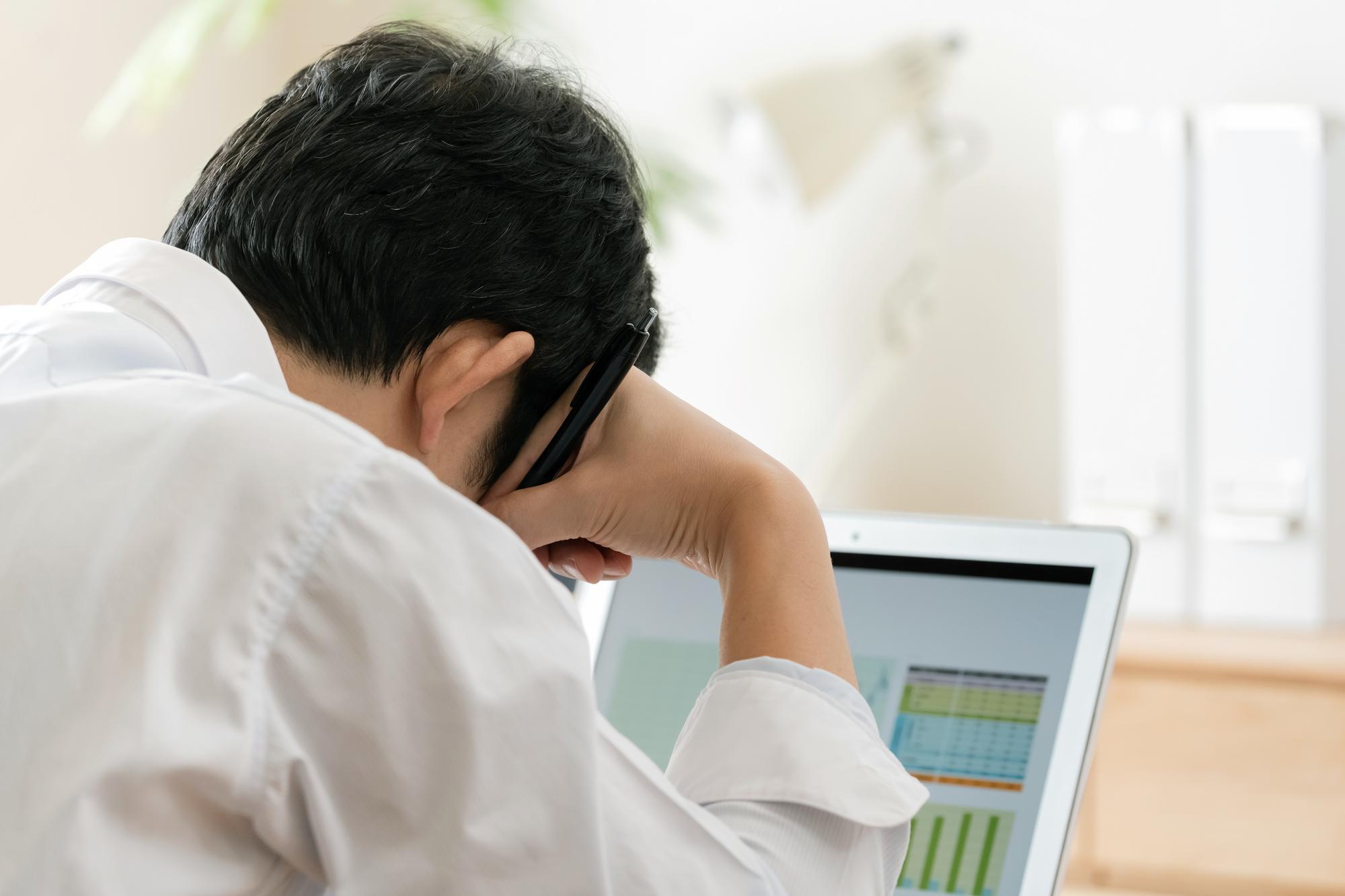Webカメラ内蔵のノートPCに向かって頭を抱える男性ビジネスパーソンの後ろ姿