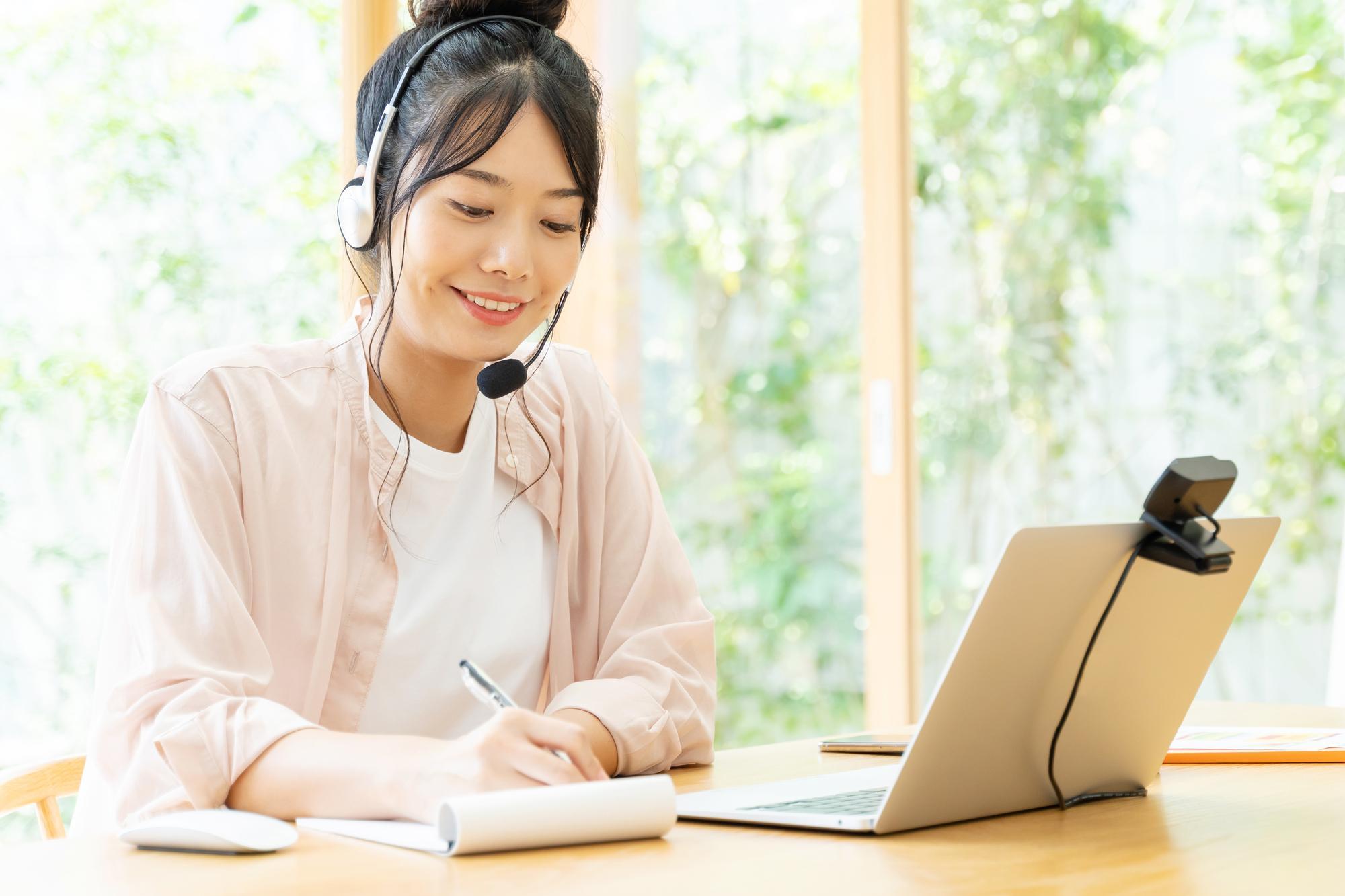 ノートPCの前でヘッドセットを着けてメモを取る若い女性