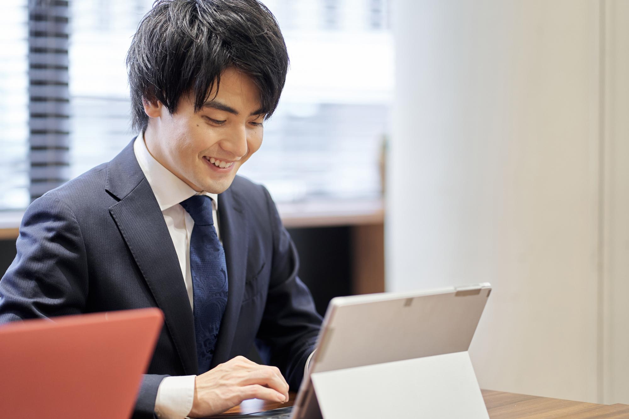 卓上に立てたタブレット型PCのキーボードを笑顔で操作する、スーツ姿の若い男性