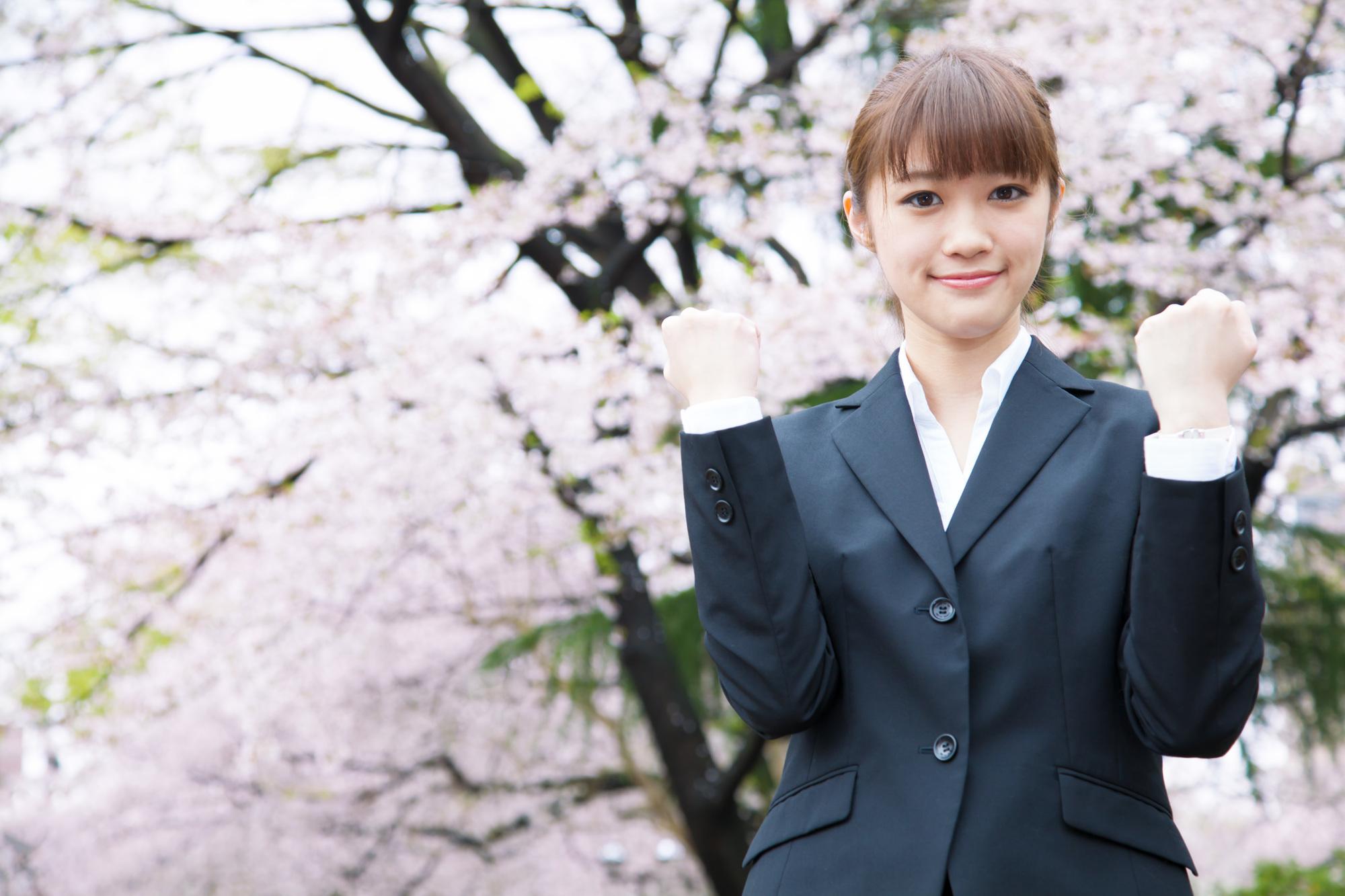 満開の桜をバックに、ガッツポーズをしてほほ笑むスーツ姿の若い女性