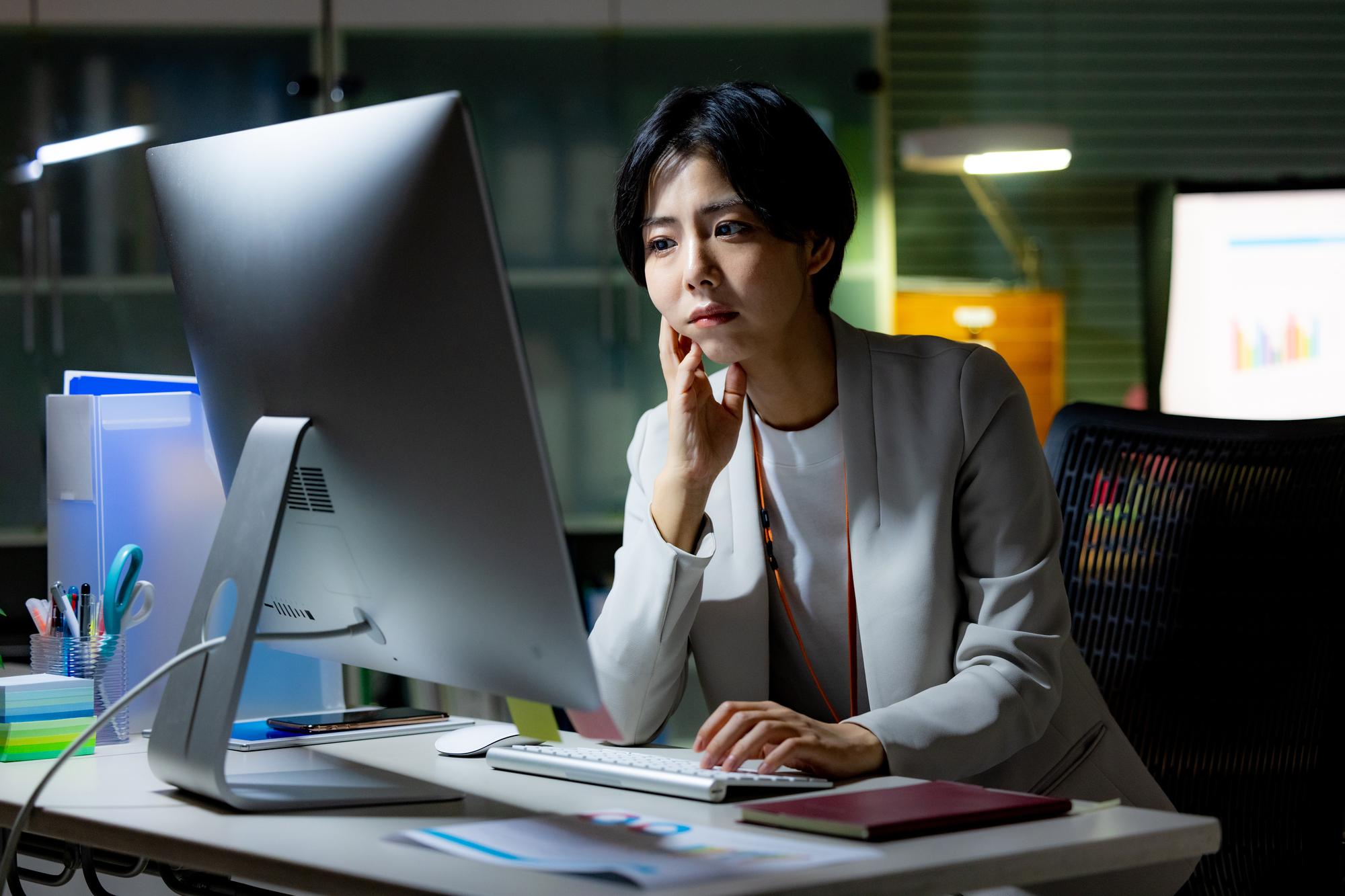 暗いオフィス内で、デスクトップPCに向かって片肘をつき悩ましげな表情を浮かべる女性