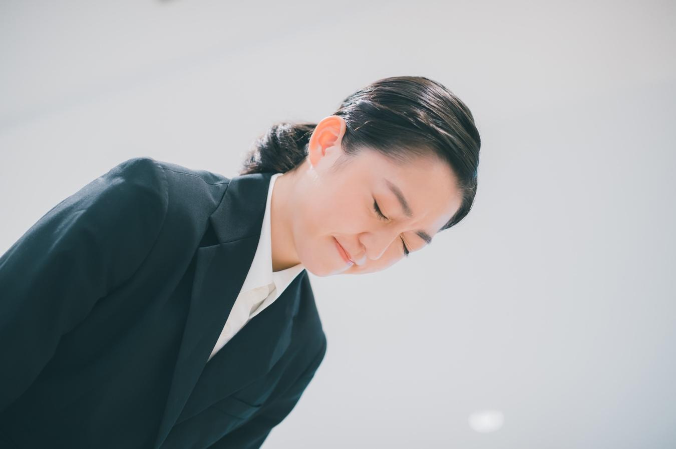 顔をしかめながら頭を下げているスーツ姿の女性