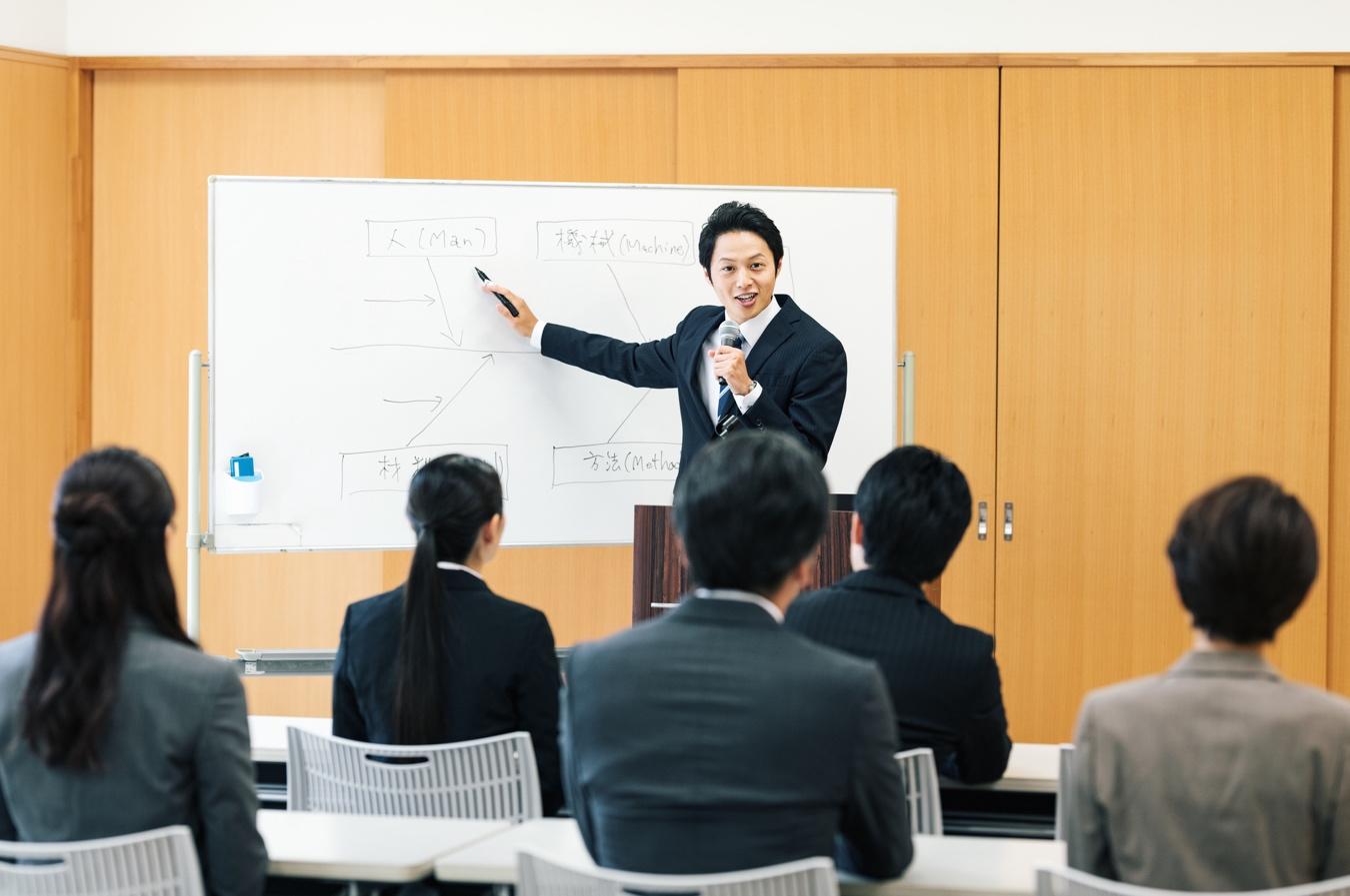 広い講義室で多数の社員を前にホワイトボードを用いて解説している男性の様子