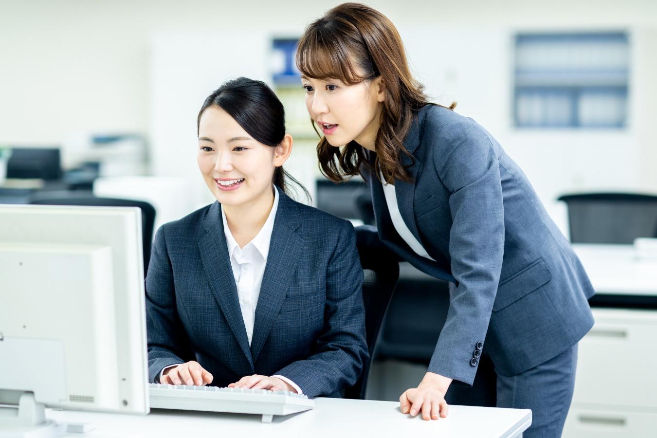 デスクトップパソコンを笑顔で操作する若い女性と、画面をのぞき込む女性