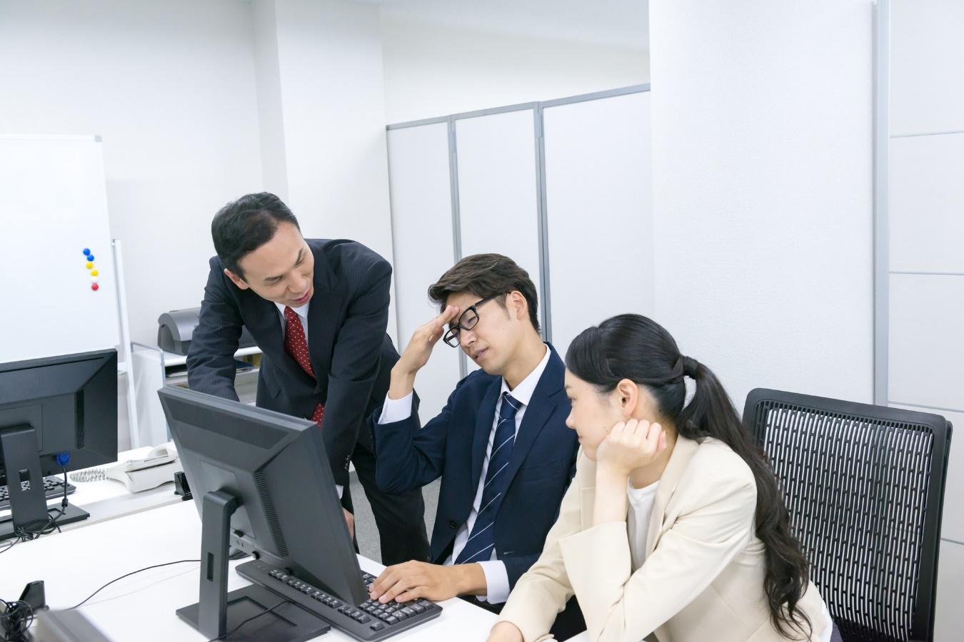 オフィス内で、ひとつのデスクトップPCを前に思い悩む男女3名