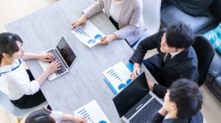 資料とノートPCを広げた会議用テーブルを囲み相談する5人男女
